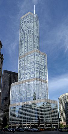 A tall silver skyscraper sits at a jog in the river beyond a bridge. The river and other along its banks buildings are in the foreground.