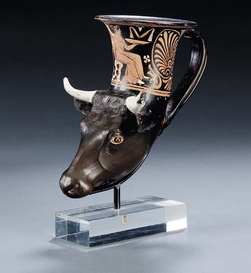 AN APULIAN RED-FIGURED RHYTON Circa 350-340 B.C. Molded in the form of a bull's head, black-glazed with added white for the horns, the ridged poll with incised details, the eyes reserved, the iris and pupil in black outlined in white, the bowl with a winged Nike seated on a rock, a phiale in her outstretched left hand, palmette complexes on either side of the strap handle, a band of dotted egg on the flaring rim 7½in. (19.1cm.) long