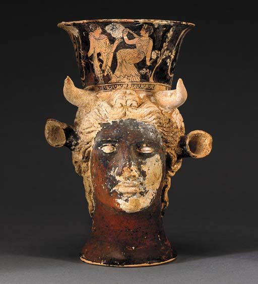 AN ATTIC RED-FIGURED FIGURAL MUG Circa 430-420 B.C. In the form of the head of Io, the human female head with bovine ears emerging from beneath her wavy locks, which are parted at the center & tied in a top knot, with short curving horns protruding forth from the top of her head; vessel neck with & encounter between Hermes & a woman, perhaps Io, the god walking left but looking back, his caduceus in his right hand, wearing a mantle tied at his neck & high boots... 7 7/8” H